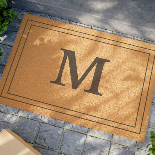 A natural coir doormat with a large dark brown letter 'M' in the center, bordered by a thin dark frame, placed on a gray stone walkway.