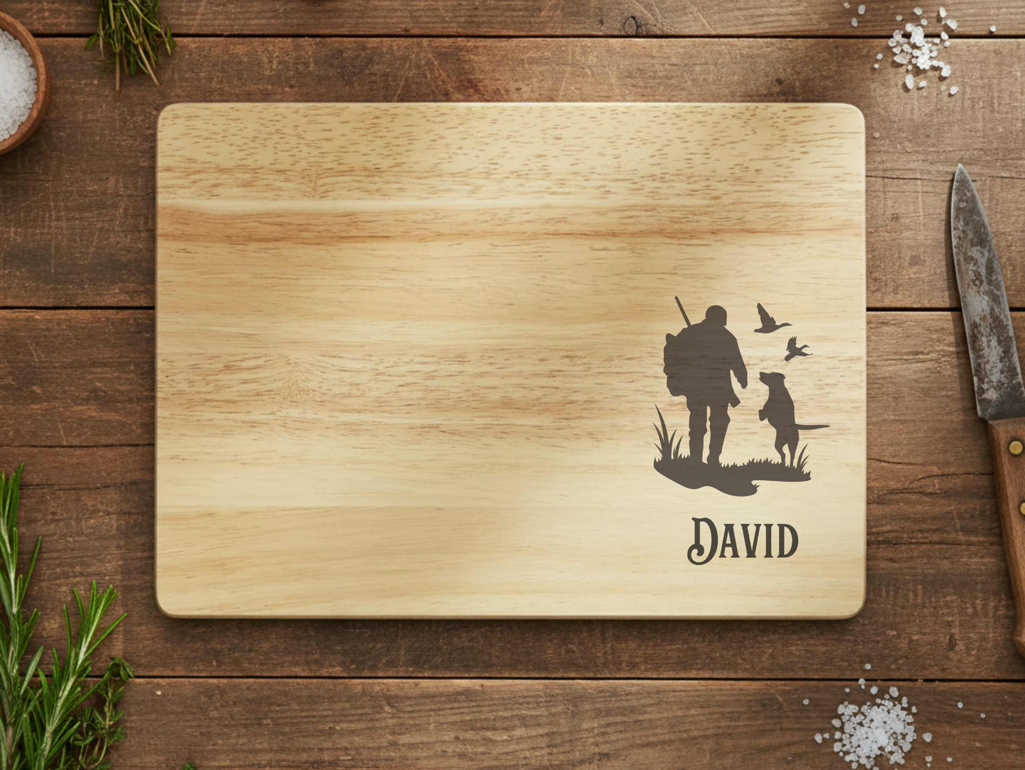 Wooden cutting board with engraved design of a hunter, dog, and birds, along with the name 'David', on a wooden surface.