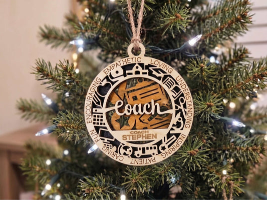 A personalized wooden Christmas ornament shaped like a circular train station map, hanging on a decorated Christmas tree. The ornament features laser-cut illustrations of a train station and a train, with the word 'Coach' and the name 'STEPHEN' in the center.