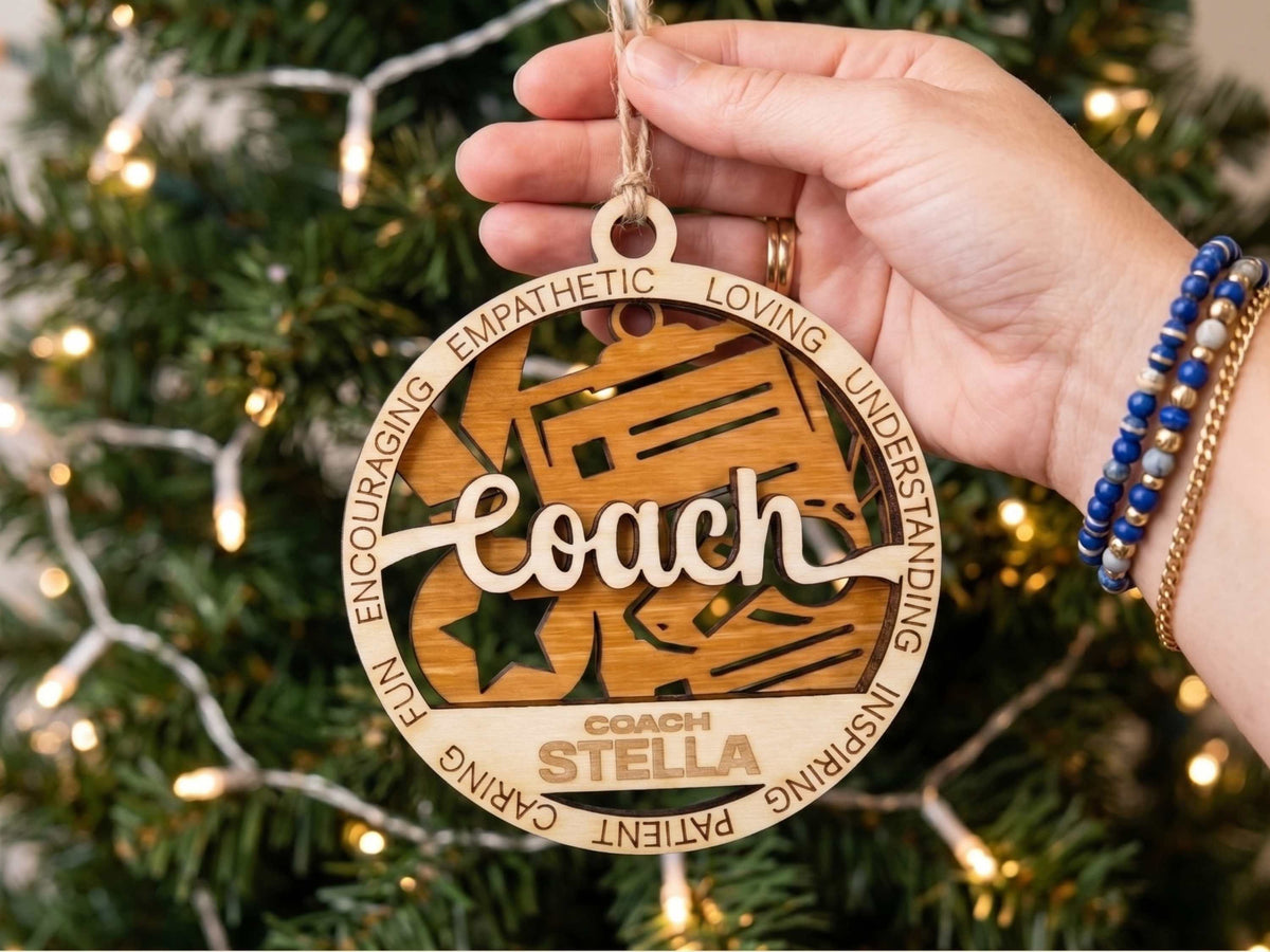 A hand holds a personalized wooden Christmas ornament shaped like a football helmet, engraved with the name 'Coach Stella' and surrounded by positive qualities, set against a blurred Christmas tree with warm lights.