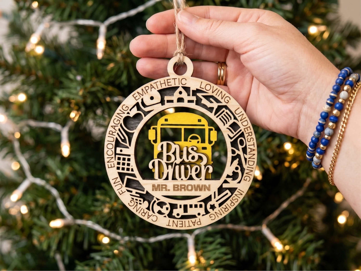 A hand holds a round wooden Christmas ornament shaped like a bus driver badge, featuring a yellow bus illustration and the words 'Bus Driver' and 'Mr. Brown', with a blurred Christmas tree in the background.