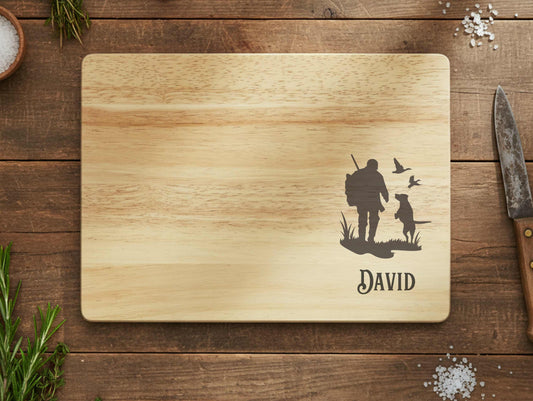 Wooden cutting board with engraved design of a hunter, dog, and birds, along with the name 'David', on a wooden surface.
