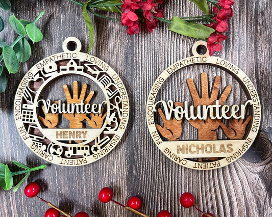 Two personalized wooden Christmas ornaments displayed on a dark wood background with red berries and green leaves. One ornament is engraved with 'Volunteer HENRY' and the other with 'Volunteer NICHOLAS', both featuring laser-cut hand graphics and service icons around the border.